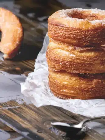 Stapel von drei Cronuts, neben einem halb gegessenen Cronut auf einem rustikalen Holzbrett.