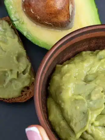 Nahaufnahme von Avocado-Creme in einer Schüssel, halber Avocado und mit Avocado-Creme bestrichenem Brot.