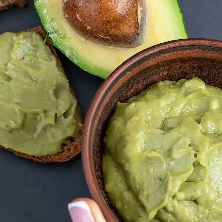 Nahaufnahme von Avocado-Creme in einer Schüssel, halber Avocado und mit Avocado-Creme bestrichenem Brot.