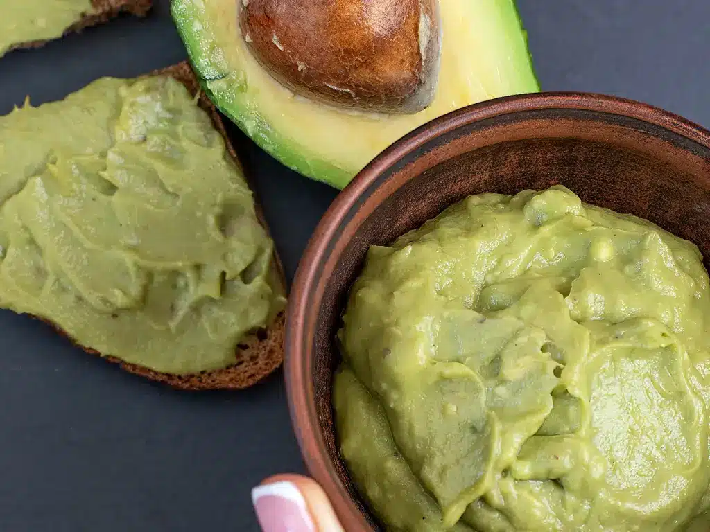 Nahaufnahme von Avocado-Creme in einer Schüssel, halber Avocado und mit Avocado-Creme bestrichenem Brot.