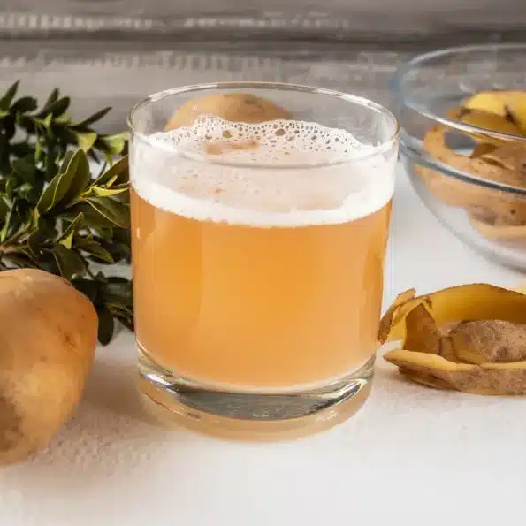 Glas mit frischem Kartoffelsaft, umgeben von Kartoffeln und Kartoffelschalen auf einem weißen Tisch.