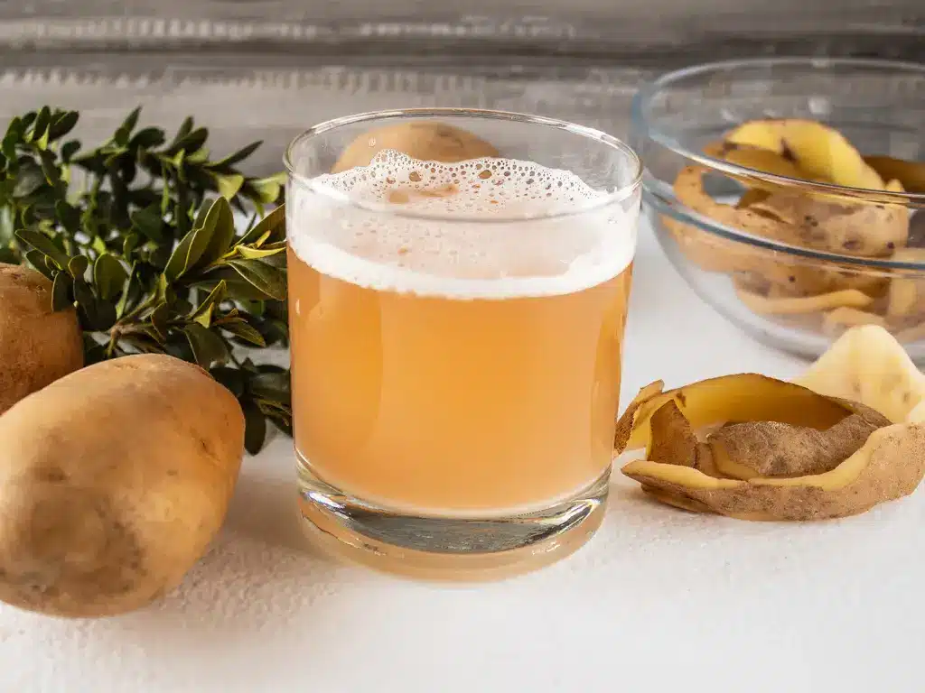 Glas mit frischem Kartoffelsaft, umgeben von Kartoffeln und Kartoffelschalen auf einem weißen Tisch.