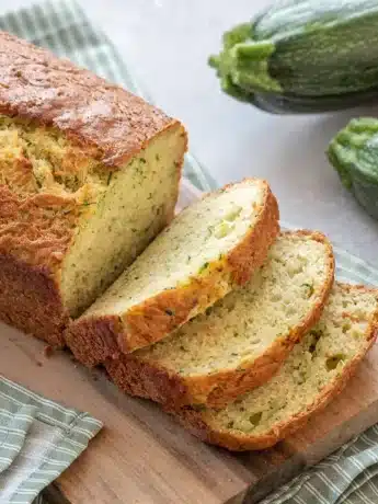 Ein frisch gebackenes Zucchinibrot, das auf einem Holzbrett liegt, mit mehreren geschnittenen Scheiben und Zucchini im Hintergrund.