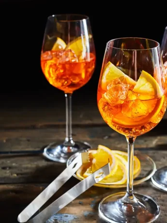 Drei Gläser Aperol Spritz mit Eis und Orangenscheiben auf einem rustikalen Holztisch