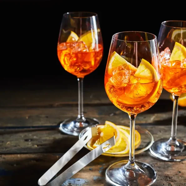 Drei Gläser Aperol Spritz mit Eis und Orangenscheiben auf einem rustikalen Holztisch