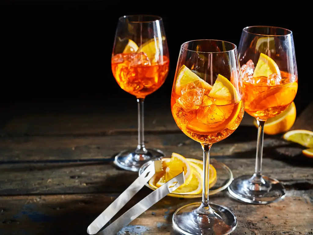 Drei Gläser Aperol Spritz mit Eis und Orangenscheiben auf einem rustikalen Holztisch