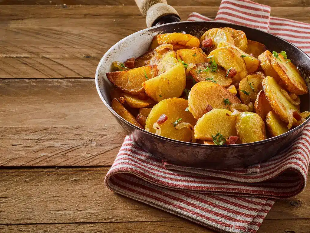 Eine Pfanne mit knusprigen Bratkartoffeln, gewürzt mit Speckstückchen und frischen Kräutern, auf einem rot-weißen Geschirrtuch.