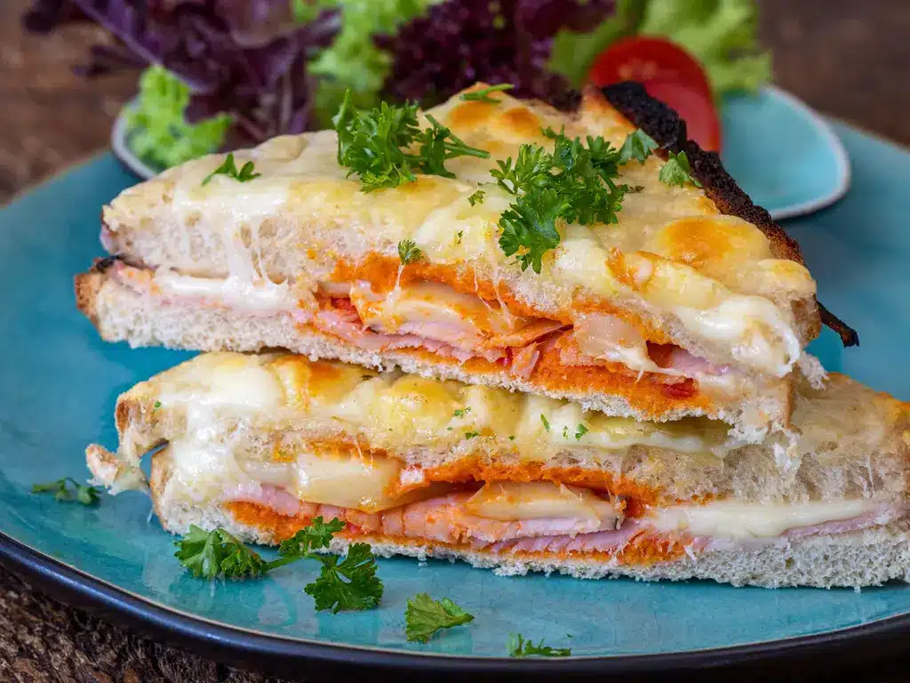 Gegrilltes Käsesandwich mit Schinken, überbacken mit Käse und garniert mit frischer Petersilie auf einem blauen Teller