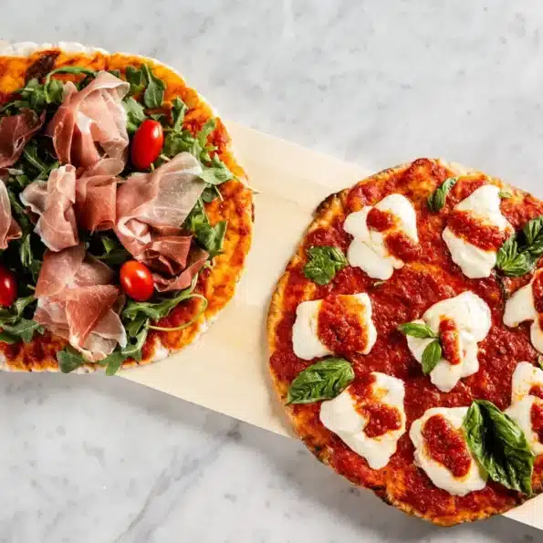 Zwei Pizzen mit glutenfreiem Pizzateig: eine mit Schinken, Rucola und Kirschtomaten und die andere mit Mozzarella und Basilikum