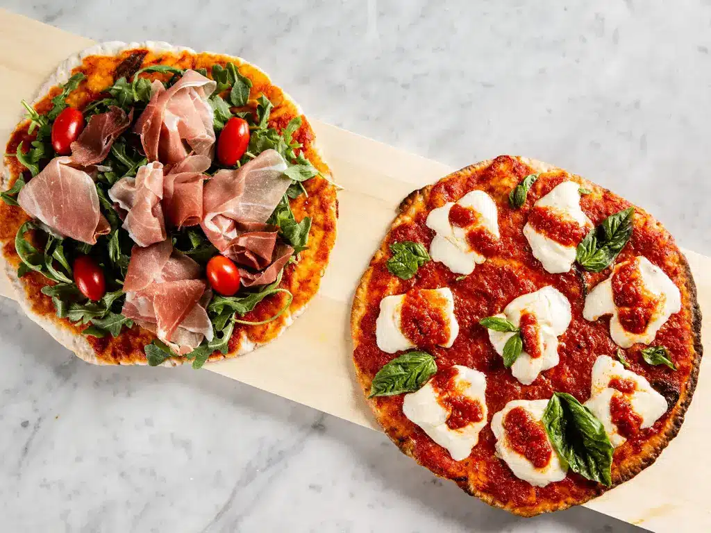 Zwei Pizzen mit glutenfreiem Pizzateig: eine mit Schinken, Rucola und Kirschtomaten und die andere mit Mozzarella und Basilikum