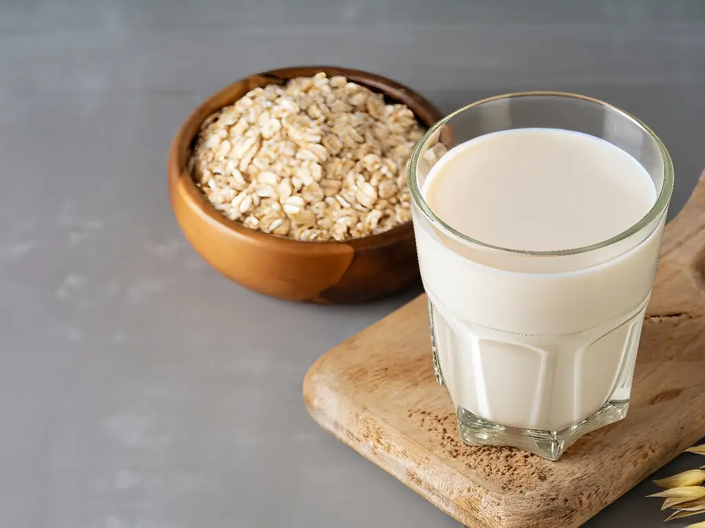 Ein Glas frische Hafermilch auf einem Holzbrett, daneben eine Schale mit Haferflocken.