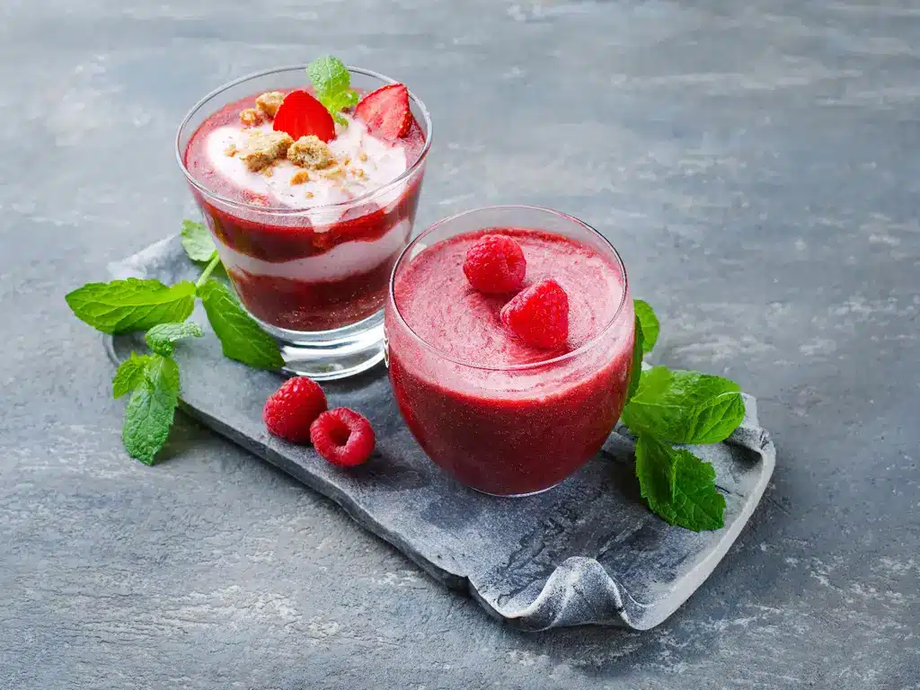 Zwei Gläser mit Himbeer- und Erdbeer-Smoothies, garniert mit frischen Beeren und Minzblättern, auf einem grauen Tablett.