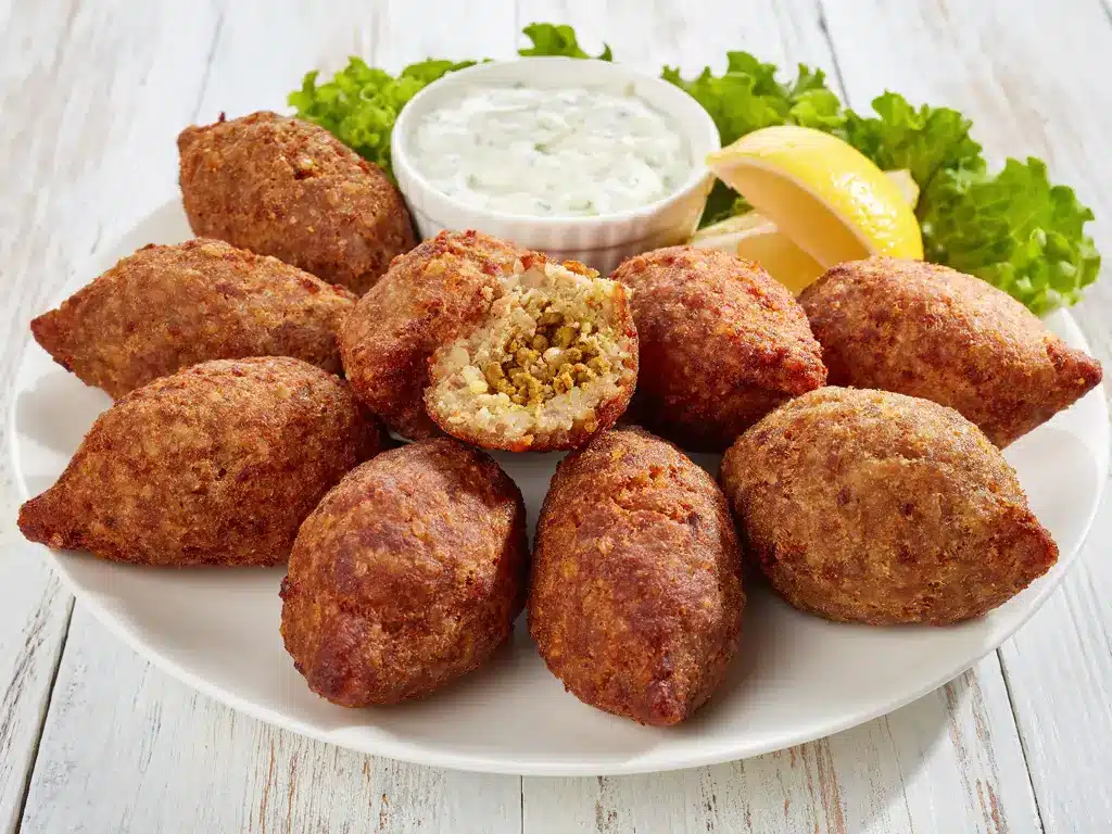 Ein Teller mit mehreren gebratenen Kibbeh, einer geöffnet, um die Füllung zu zeigen, daneben eine Schale mit Joghurt-Dip und Zitronenscheiben.