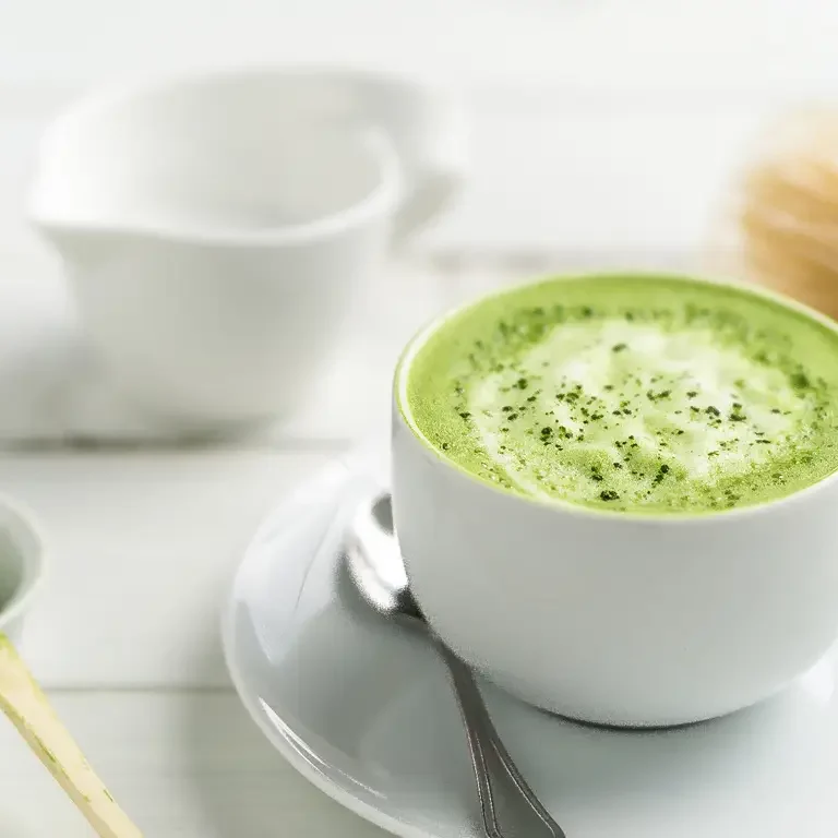 Eine Tasse cremiger Matcha Latte mit Schaum auf einem weißen Unterteller, daneben ein Matcha-Besen und ein Löffel mit Matcha-Pulver.