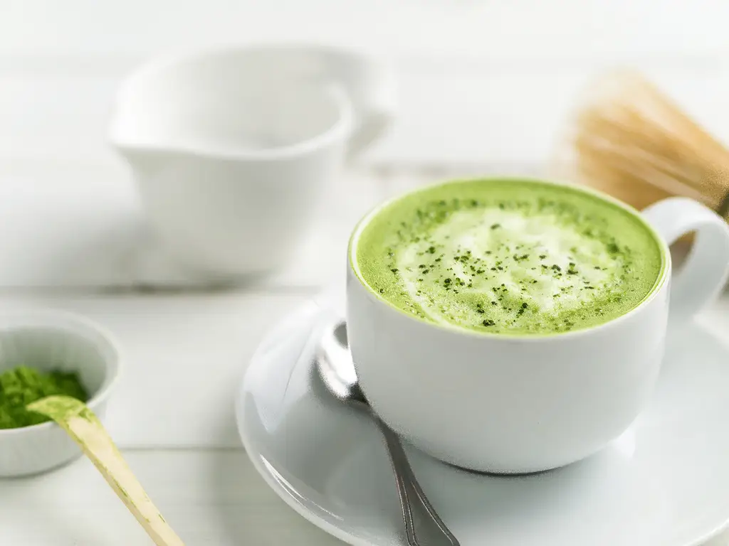 Eine Tasse cremiger Matcha Latte mit Schaum auf einem weißen Unterteller, daneben ein Matcha-Besen und ein Löffel mit Matcha-Pulver.