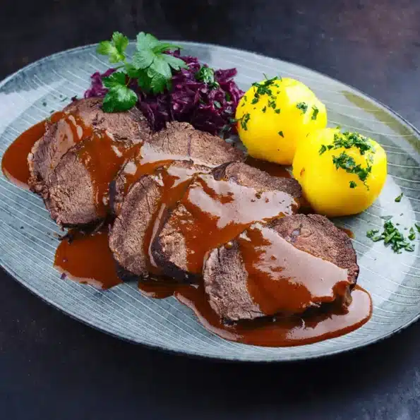 Ein Teller mit zartem Rinderbraten in Bratensauce, serviert mit Kartoffelklößen und Rotkohl, garniert mit frischen Kräutern.