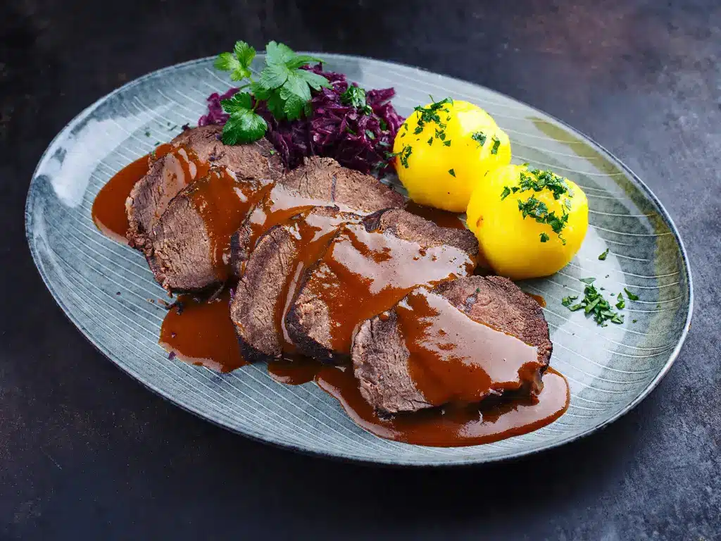 Ein Teller mit zartem Rinderbraten in Bratensauce, serviert mit Kartoffelklößen und Rotkohl, garniert mit frischen Kräutern.