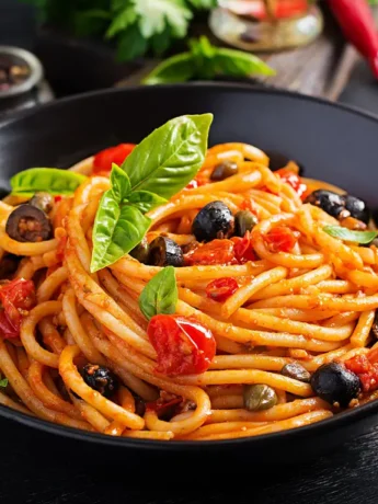 Eine Schale mit Spaghetti alla Puttanesca, garniert mit frischem Basilikum, Tomaten, Oliven und Kapern.