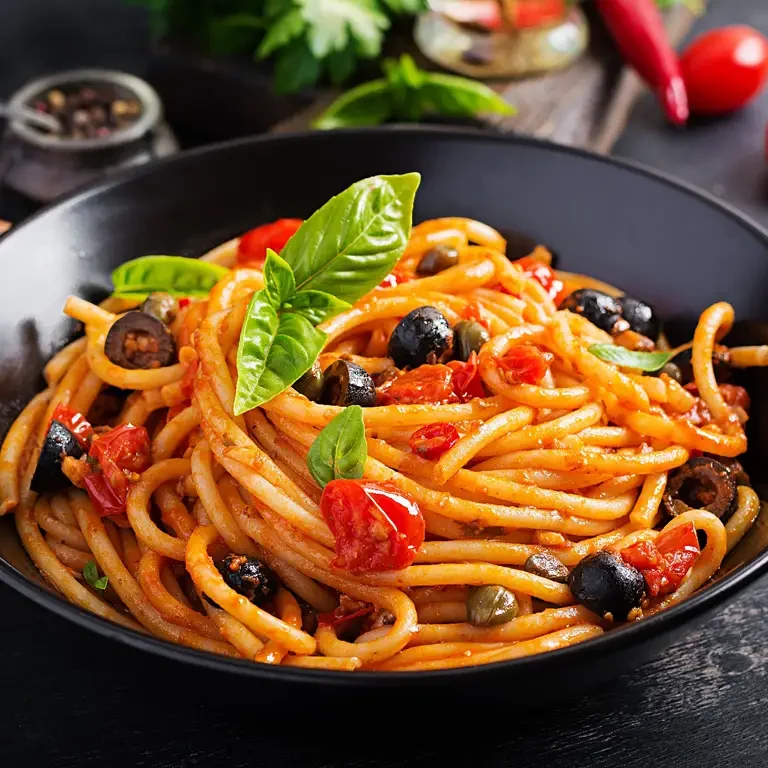 Eine Schale mit Spaghetti alla Puttanesca, garniert mit frischem Basilikum, Tomaten, Oliven und Kapern.