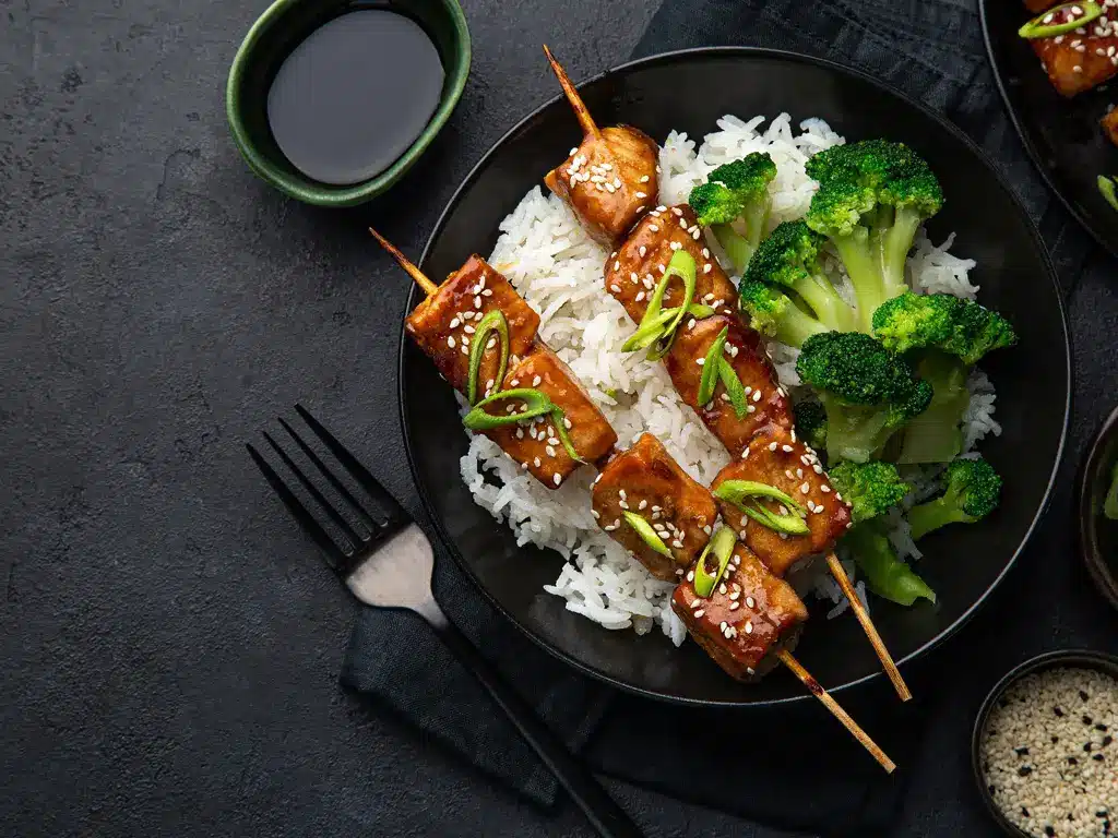 Eine Schale mit Teriyaki-Lachs-Spießen, serviert auf Reis und garniert mit Frühlingszwiebeln und Sesam, daneben Brokkoli.