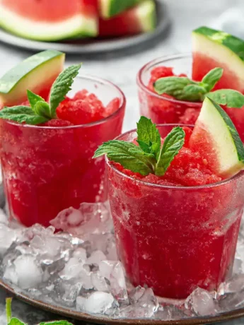 Drei Gläser mit Wassermelonen-Slush, garniert mit Minzblättern und Wassermelonenscheiben, serviert auf Eis.